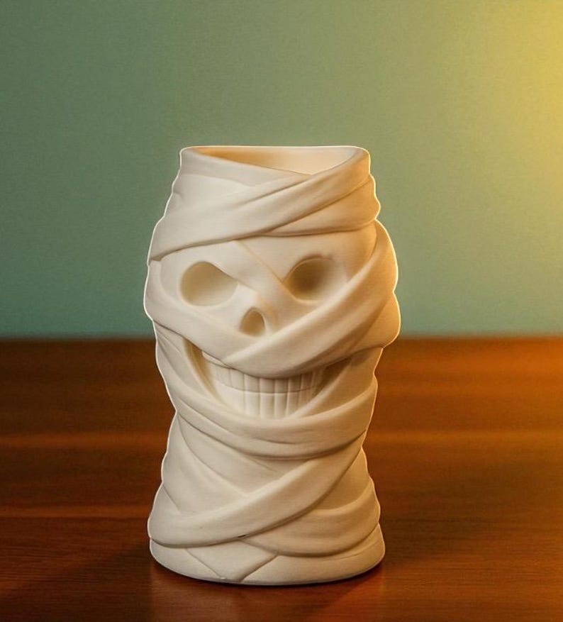 May include: A white ceramic tiki mug shaped like a mummy's head. The mug features a skull face with wrapped bandages, creating a spooky and unique design. The mug is designed for beverages and is suitable for Halloween or themed parties.