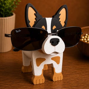 May include: A wooden dog-shaped eyeglass holder with a black nose and sunglasses. The dog is white with brown and black accents. The sunglasses are dark-tinted with the Ray-Ban logo on the left lens. The holder is on a wooden surface.
