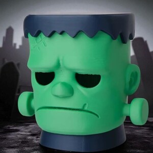 May include: A green Frankenstein-themed container with a blue top and base. The container features a detailed face with stitched scars, black eye sockets, and a furrowed brow. The background shows a silhouette of a graveyard and city.