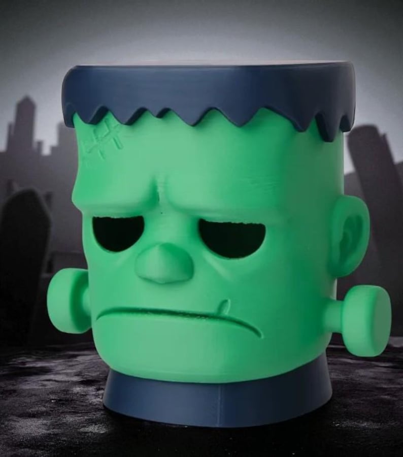 May include: A green Frankenstein-themed container with a blue top and base. The container features a detailed face with stitched scars, black eye sockets, and a furrowed brow. The background shows a silhouette of a graveyard and city.