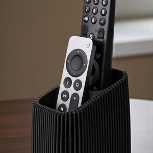 May include: A black, ribbed remote control holder containing two remotes. One is black with numbered buttons, the other silver with a circular control pad. The holder sits on a wooden surface.