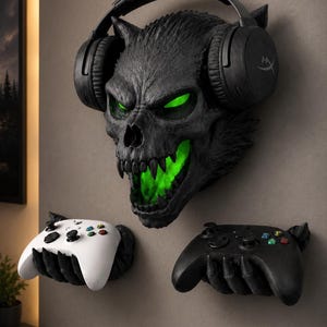 Puede incluir: Una escultura de pared con forma de cráneo de criatura negro con ojos y boca verde brillante, que lleva auriculares negros. Debajo del cráneo, dos manos negras con garras sobresalen de la pared, cada una sosteniendo un mando de juego, uno blanco y otro negro.