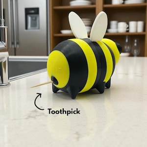 May include: A whimsical bee-shaped toothpick holder. The holder is black and yellow, with a striped design and white wings. A wooden toothpick is inserted into the holder. The word "Toothpick" is written below the holder.