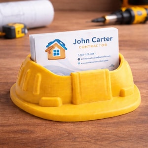 May include: A yellow construction helmet-shaped business card holder, holding white business cards. The cards display the name "John Carter, Contractor". The holder is on a wooden surface. The design is 3D-printed.