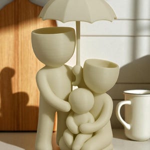 May include: A cream-colored family figurine, featuring a parent figure holding an umbrella over a family of three. The figurine has a minimalist design with open-top heads, possibly for plants or small items. A mug is visible in the background.