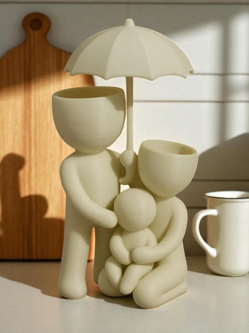 May include: A cream-colored family figurine, featuring a parent figure holding an umbrella over a family of three. The figurine has a minimalist design with open-top heads, possibly for plants or small items. A mug is visible in the background.