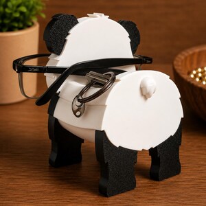 May include: A white and black panda-shaped eyeglasses holder with black eyeglasses and a key ring. The holder has black ears, feet, and a tail. The eyeglasses are black with the brand name "Zeri" on the left arm. The holder is on a wooden surface.
