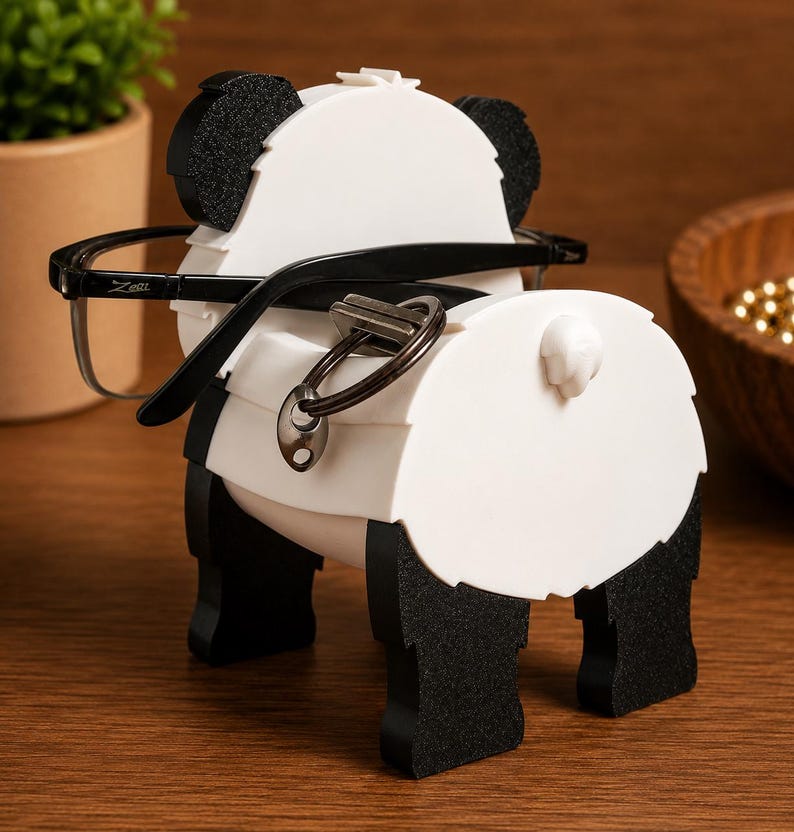 May include: A white and black panda-shaped eyeglasses holder with black eyeglasses and a key ring. The holder has black ears, feet, and a tail. The eyeglasses are black with the brand name "Zeri" on the left arm. The holder is on a wooden surface.