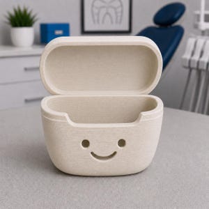 May include: Beige, open container with a smiling face design. The container has a rectangular shape with rounded edges and a textured surface. The lid is open, revealing the interior. The background includes a dental chair and a framed dental chart.
