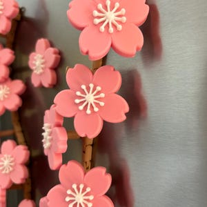 May include: Pink cherry blossom magnets with white centers are arranged on a brown stem. The magnets are attached to a silver surface. The blossoms are in full bloom, with multiple magnets visible.