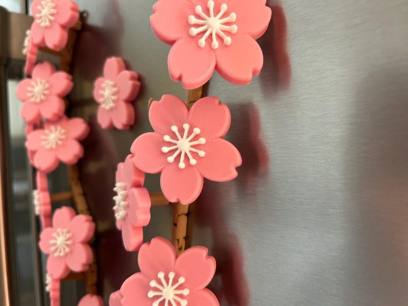 May include: Pink cherry blossom magnets with white centers are arranged on a brown stem. The magnets are attached to a silver surface. The blossoms are in full bloom, with multiple magnets visible.