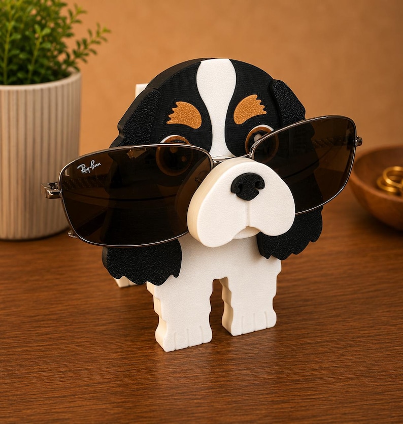 Pu&ograve; includere: Un originale portaocchiali a forma di cane, nero, bianco e marrone. Il supporto &egrave; progettato per contenere un paio di occhiali da sole, con le lenti appoggiate sul muso del cane. Gli occhiali da sole hanno una montatura argentata e lenti scure.