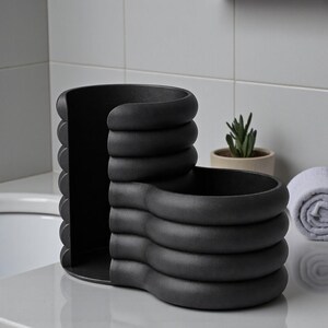 Pode incluir: Um organizador de banheiro preto fosco e moderno com um design exclusivo em camadas. O organizador apresenta um compartimento curvo com a parte superior aberta e uma ranhura vertical, perfeito para guardar produtos de higiene pessoal ou acess&oacute;rios. Uma pequena suculenta em um vaso e uma toalha branca enrolada est&atilde;o ao fundo.