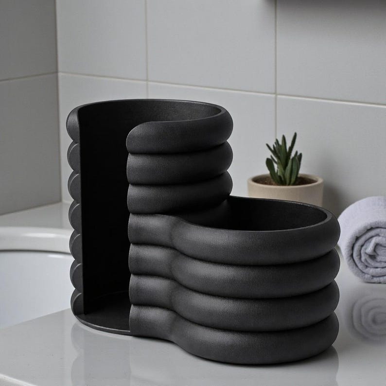 Pode incluir: Um organizador de banheiro preto fosco e moderno com um design exclusivo em camadas. O organizador apresenta um compartimento curvo com a parte superior aberta e uma ranhura vertical, perfeito para guardar produtos de higiene pessoal ou acess&oacute;rios. Uma pequena suculenta em um vaso e uma toalha branca enrolada est&atilde;o ao fundo.
