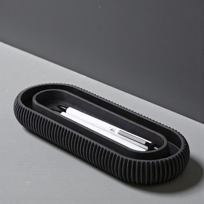 May include: A black, oval-shaped desk organizer with a ribbed exterior. Inside, a smaller oval holds two white pens with black accents. The organizer sits on a gray surface, offering a modern, minimalist aesthetic.