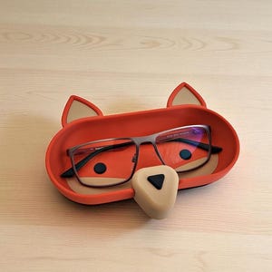 May include: A red fox-shaped glasses holder with a beige nose and ears. The holder contains a pair of eyeglasses with dark frames. The fox face has black eyes and is resting on a light wooden surface.