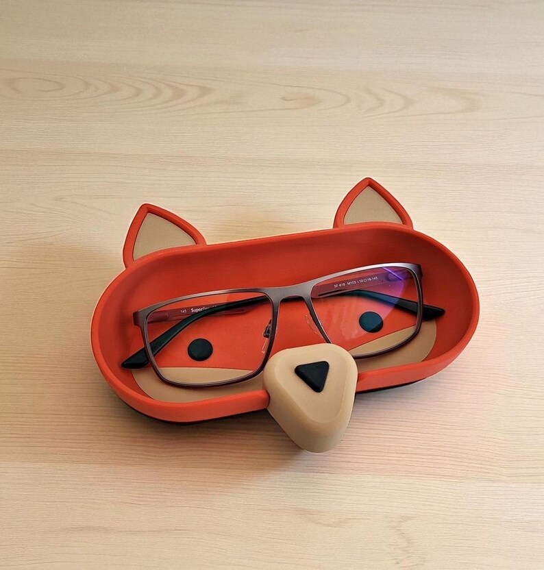 May include: A red fox-shaped glasses holder with a beige nose and ears. The holder contains a pair of eyeglasses with dark frames. The fox face has black eyes and is resting on a light wooden surface.