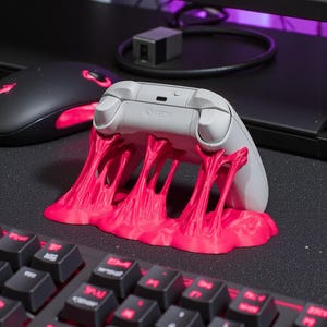 May include: A grey Xbox controller sits on a bright pink stand, designed to look like melting slime. The controller is above a black keyboard with red illuminated keys. A black mouse with red accents is also visible.