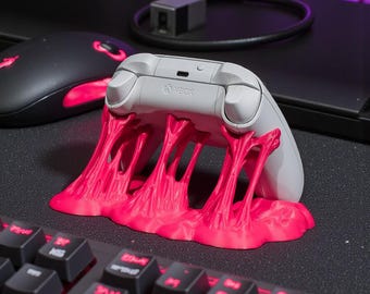 May include: A gray Xbox controller rests on a vibrant pink stand, resembling melting slime. The controller is positioned above a black keyboard with red illuminated keys. A black mouse with red accents is visible.