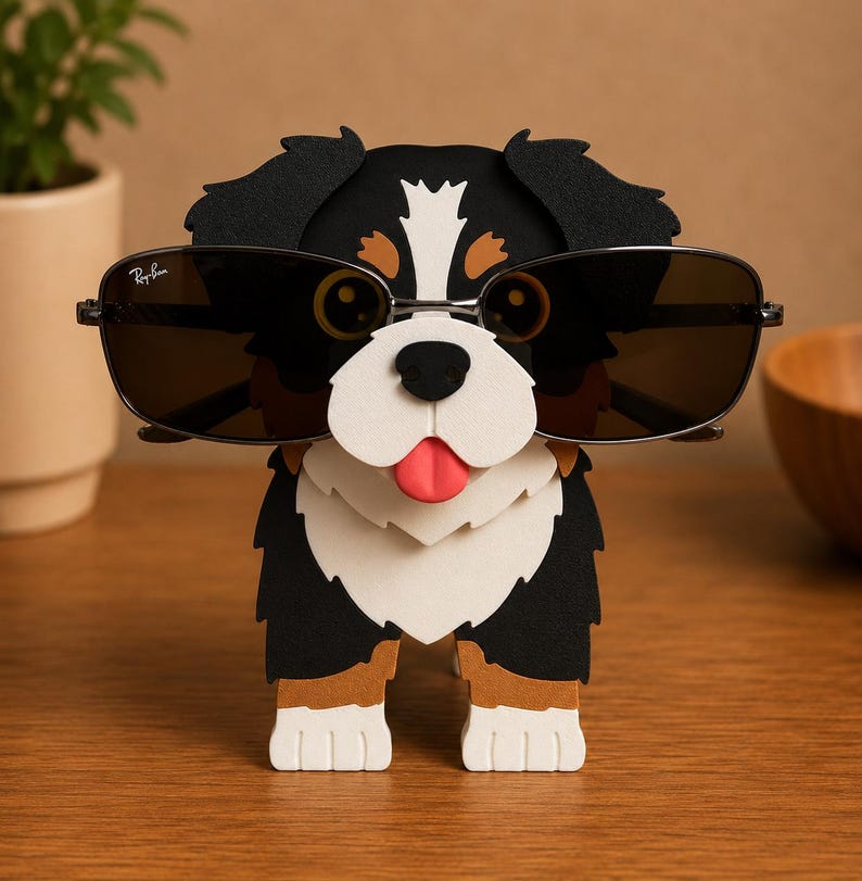 May include: A decorative dog-shaped eyeglass holder. The holder is black, white, and brown, with a red tongue. It is wearing sunglasses with the Ray-Ban logo. The holder is made of layered materials and sits on a wooden surface.