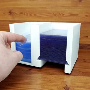 May include: A white, two-compartment card holder with a stack of blue card sleeves. A hand is pulling a sleeve from the left compartment. The holder sits on a wooden surface.