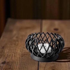 May include: A black metal candle holder with a geometric diamond pattern. The candle holder has a round base and holds a white tea light candle. The holder sits on a wooden surface.