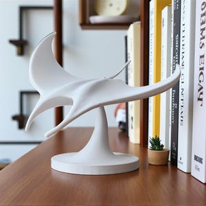 May include: A white, abstract sculpture resembling a manta ray, set on a wooden surface. The sculpture features a smooth, flowing design and a circular base. Books and a small potted plant are visible in the background.
