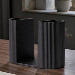May include: A black desk tidy with a distinctive design. It features a rectangular section and a cylindrical section with vertical ridges. It is placed on a wooden surface.