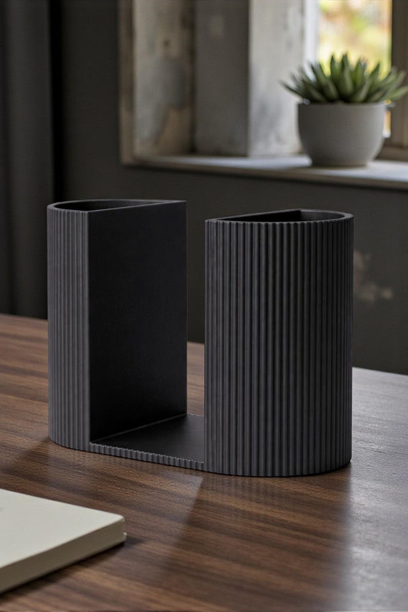 May include: A black desk tidy with a distinctive design. It features a rectangular section and a cylindrical section with vertical ridges. It is placed on a wooden surface.