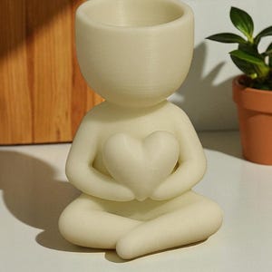 May include: A cream-colored, human-shaped planter holding a heart. The planter is in a seated position with its legs crossed. The top of the head is a small, open bowl for planting. The planter is made of a smooth, matte material.