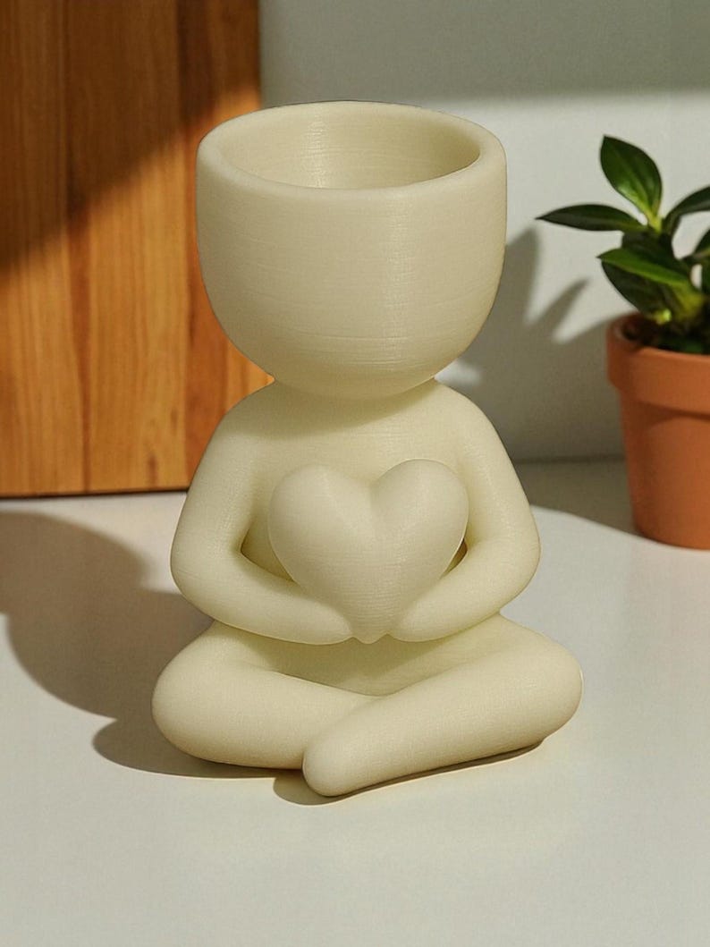 May include: A cream-colored, human-shaped planter holding a heart. The planter is in a seated position with its legs crossed. The top of the head is a small, open bowl for planting. The planter is made of a smooth, matte material.