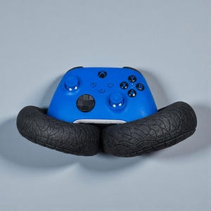 May include: A blue Xbox controller is cradled by two black, textured tire-shaped holders. The controller has the Xbox logo and various buttons. The background is a solid light gray.