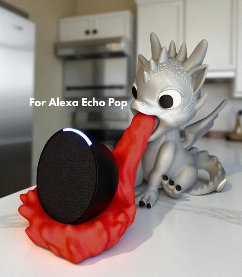 May include: A silver dragon-shaped speaker holder with a red tongue holding a black speaker. The text "For Alexa Echo Pop" is visible. The speaker has a black mesh front and a white light.
