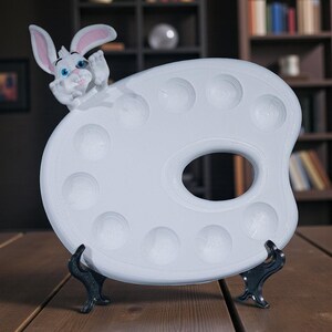 May include: A white ceramic artist's palette with a cartoon bunny figure. The palette has multiple circular wells and a thumb hole. The bunny has pink-lined ears and blue eyes. The palette is supported by a black stand.
