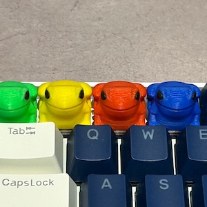 May include: Four frog-shaped keycaps in green, yellow, red, and blue, sitting on a keyboard with the keys "Tab", "CapsLock", "Q", "W", "E", "A", "S", "D", and "4$", "F4".