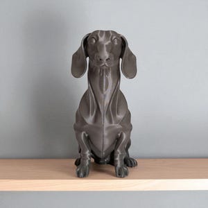 May include: A dark gray dachshund dog statue sitting on a light wood shelf. The dog has long ears and a long body, with its front paws resting on the shelf. The statue is a solid color and has a smooth finish.