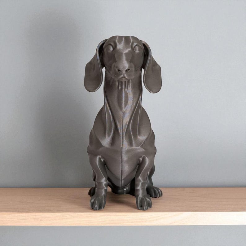 May include: A dark gray dachshund dog statue sitting on a light wood shelf. The dog has long ears and a long body, with its front paws resting on the shelf. The statue is a solid color and has a smooth finish.