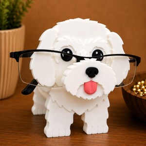 May include: A white, 3D-printed dog-shaped eyeglasses holder with black eyes, a black nose, and a pink tongue. The dog is wearing black-framed glasses. The holder is on a wooden surface.