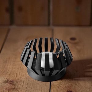 Puede incluir: Un portavelas de metal negro con un dise&ntilde;o geom&eacute;trico. El soporte tiene una base circular y una estructura en forma de jaula con barras verticales. Una vela de t&eacute; plateada se encuentra dentro del portavelas. El portavelas est&aacute; sobre una superficie de madera.