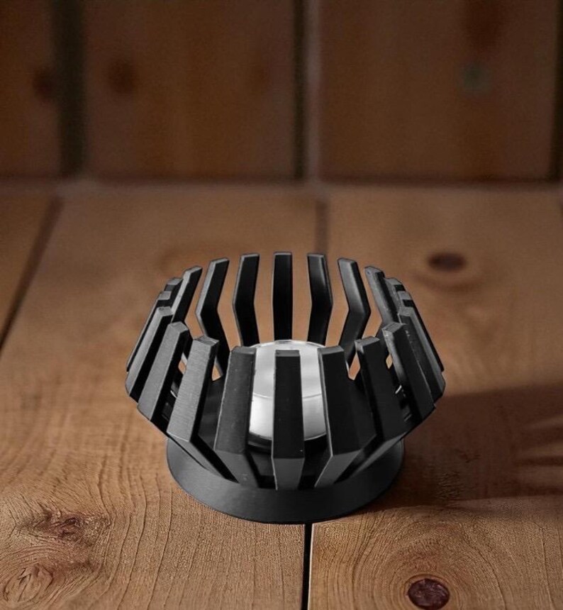 Puede incluir: Un portavelas de metal negro con un dise&ntilde;o geom&eacute;trico. El soporte tiene una base circular y una estructura en forma de jaula con barras verticales. Una vela de t&eacute; plateada se encuentra dentro del portavelas. El portavelas est&aacute; sobre una superficie de madera.