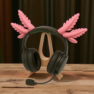 Puede incluir: Auriculares negros de diadema con micr&oacute;fono, adornados con detalles decorativos rosas en forma de plumas. Los auriculares descansan sobre un soporte de madera, sobre un fondo de madera. El dise&ntilde;o es caprichoso y creativo.
