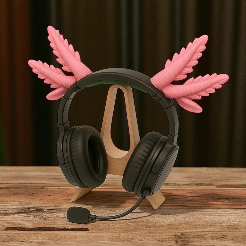 Puede incluir: Auriculares negros de diadema con micr&oacute;fono, adornados con detalles decorativos rosas en forma de plumas. Los auriculares descansan sobre un soporte de madera, sobre un fondo de madera. El dise&ntilde;o es caprichoso y creativo.