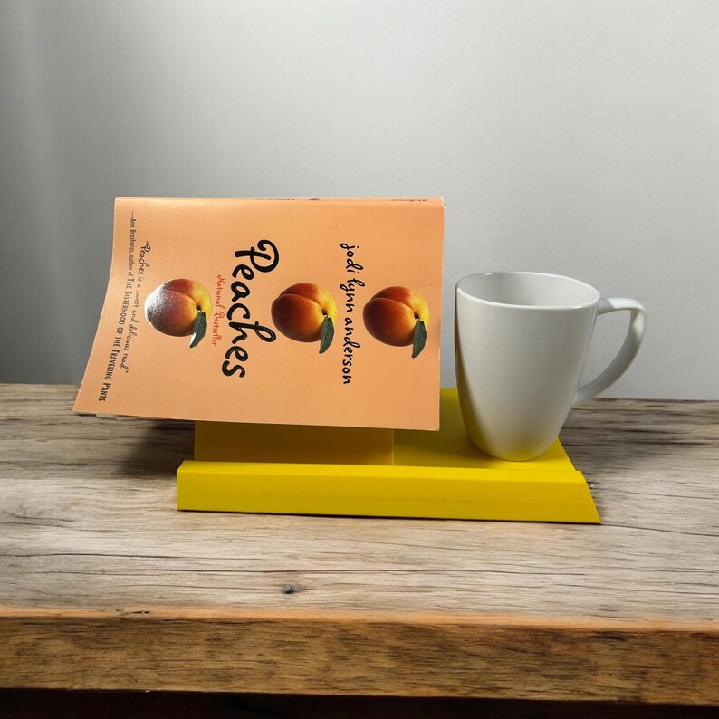 May include: A book titled "Peaches" by Jodi Lynn Anderson, featuring peach illustrations, rests on a yellow stand next to a white mug. The book's cover is peach-colored.