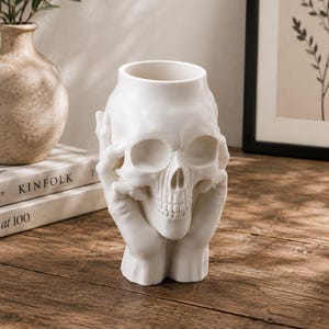 May include: A white ceramic skull-shaped vase with hands cradling the skull. The vase has a hollow interior for holding flowers or other decorative items. The skull's features are detailed, and the hands add a unique touch to the design.