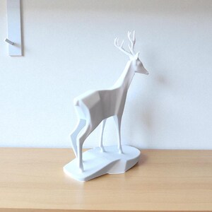 May include: A white geometric deer sculpture with antlers, standing on a white base. The deer is positioned on a light brown wooden surface, with a white wall in the background. A white wall hook is visible on the wall.