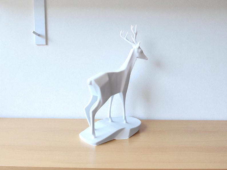 May include: A white geometric deer sculpture with antlers, standing on a white base. The deer is positioned on a light brown wooden surface, with a white wall in the background. A white wall hook is visible on the wall.