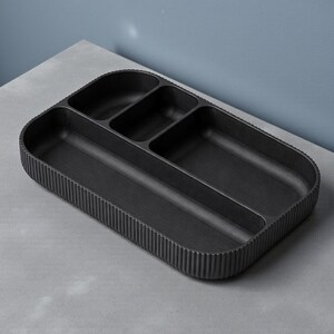 May include: A black rectangular organizer tray with multiple compartments. The tray has a ribbed exterior and a smooth interior, designed for organizing items. The tray is made of a dark material and sits on a gray surface.
