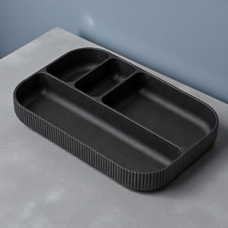 May include: A black rectangular organizer tray with multiple compartments. The tray has a ribbed exterior and a smooth interior, designed for organizing items. The tray is made of a dark material and sits on a gray surface.