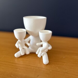 May include: A white ceramic planter shaped like a figure with two smaller figures attached. The planter has a large, open bowl for planting. The smaller figures appear to be seated, adding a whimsical touch to the design.