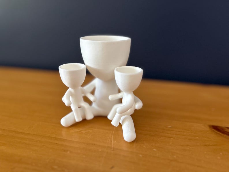 May include: A white ceramic planter shaped like a figure with two smaller figures attached. The planter has a large, open bowl for planting. The smaller figures appear to be seated, adding a whimsical touch to the design.